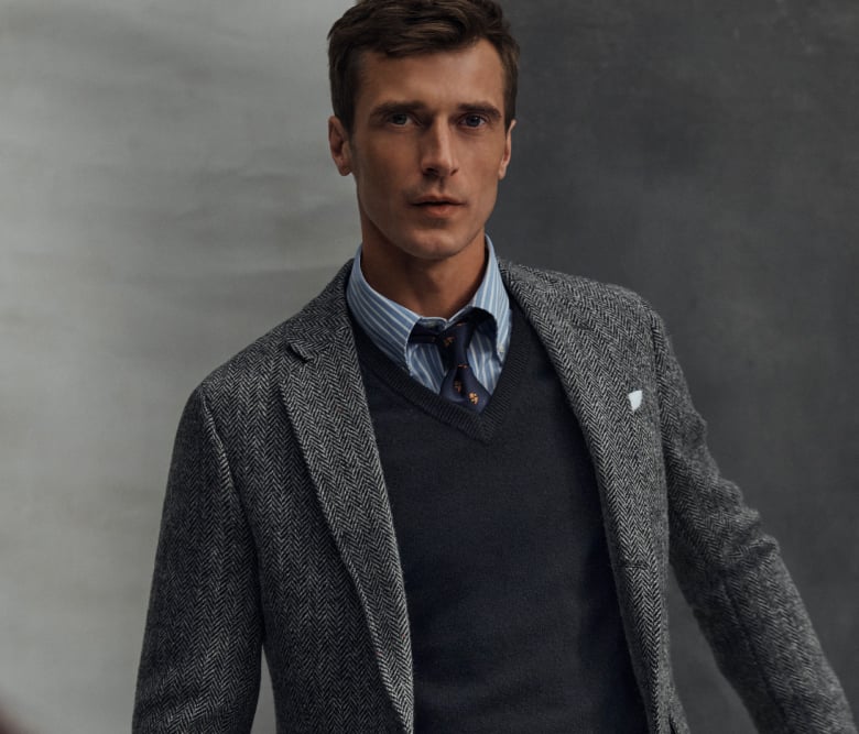 Ein Mann trägt einen grauen Blazer, der durch einen dunkelgrauen Pullover, ein blaues Hemd und eine passende Krawatte ergänzt wird