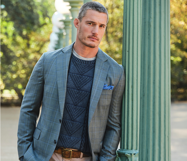 A gentleman dressed in a grey blazer, complemented by a dark grey jumper, a blue dress shirt, and a coordinating tie