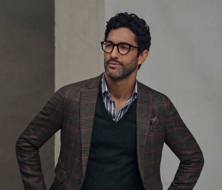 Ein Mann in einem eleganten Look mit Blazer, Pullover und Freizeithemd, ergänzt durch eine elegante schwarze Brille